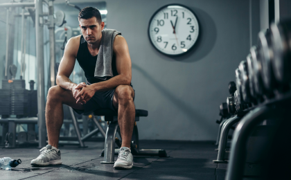 Stress und Muskelaufbau. Mann sitzt nach hartem Training erschöpft und frustriert im Fitnessstudio und symbolisiert den negativen Einfluss von Stress auf den Muskelaufbau.