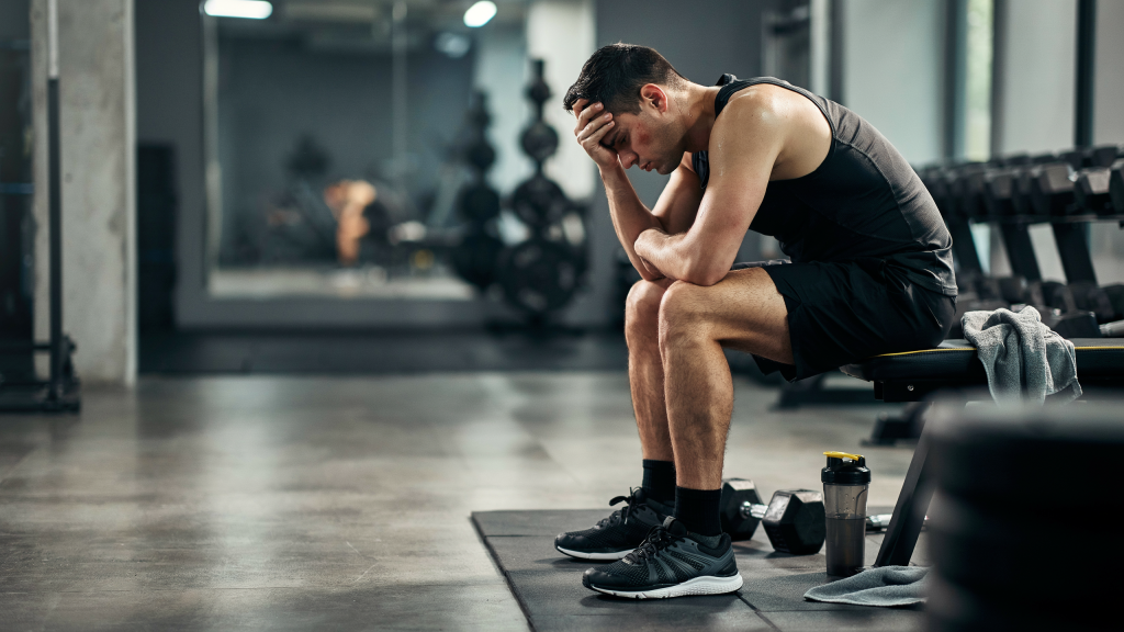 Trotz Training keine Fortschritte. Erschöpfter Sportler sitzt nach dem Training frustriert auf einer Hantelbank im Fitnessstudio, weil trotz regelmäßigem Training keine Fortschritte sichtbar sind.
