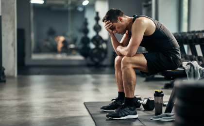 Trotz Training keine Fortschritte. Erschöpfter Sportler sitzt nach dem Training frustriert auf einer Hantelbank im Fitnessstudio, weil trotz regelmäßigem Training keine Fortschritte sichtbar sind.
